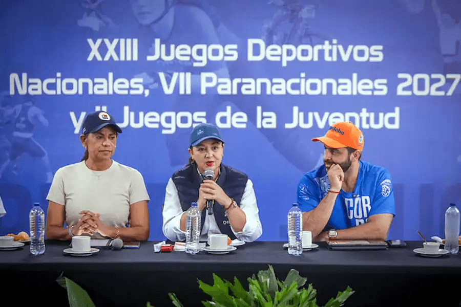 Tres voceros de los Juegos en la mesa de una rueda de prensa; la del centro habla por micrófono ante el telón del evento